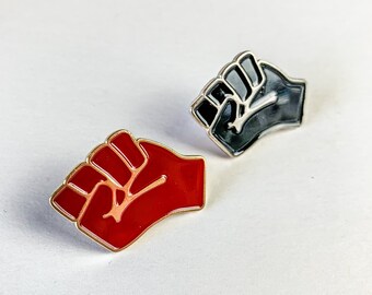 May include: Two enamel pins in the shape of a raised fist. One pin is red and the other is black.