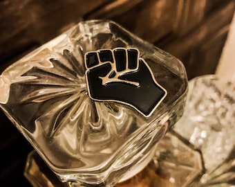 May include: A gold-colored pin with a black raised fist design. The pin is on a clear glass surface.