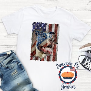 以下が含まれることがあります： アメリカ国旗のデザインが施された白いTシャツ。ピットブルの犬が描かれており、犬の顔は国旗で一部隠されています。犬の舌は出ています。画像の下には「American Pie Graphics」という文字が印刷されています。