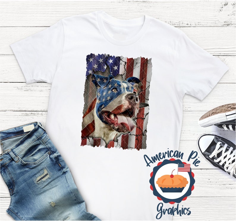 以下が含まれることがあります： アメリカ国旗のデザインが施された白いTシャツ。ピットブルの犬が描かれており、犬の顔は国旗で一部隠されています。犬の舌は出ています。画像の下には「American Pie Graphics」という文字が印刷されています。