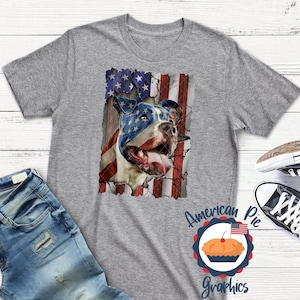 以下が含まれることがあります： アメリカ国旗のデザインが施されたグレーのTシャツ。ピットブルの犬が描かれており、犬の顔は国旗で一部隠されています。画像の下には「American Pie Graphics」というテキストが印刷されています。