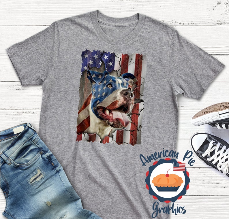 以下が含まれることがあります： アメリカ国旗のデザインが施されたグレーのTシャツ。ピットブルの犬が描かれており、犬の顔は国旗で一部隠されています。画像の下には「American Pie Graphics」というテキストが印刷されています。