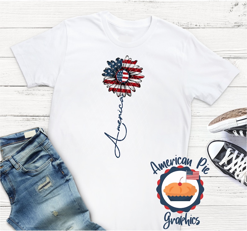 May include: White t-shirt with a red, white, and blue sunflower design and the word "America" written vertically down the stem. The design is from American Pie Graphics.