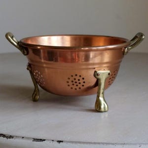 May include: A copper colander with brass handles and feet. The bowl-shaped colander has small holes for draining and is supported by three decorative brass legs. The handles are curved and attached to the rim.
