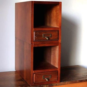 May include: A tall, wooden storage unit with a warm, reddish-brown finish. It features two open shelves and two small drawers with brass-toned label holders and pulls. The unit is designed to be stacked.