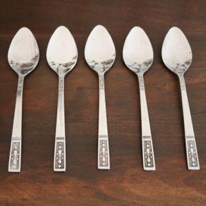 May include: Five silver spoons arranged in a row. Each spoon has an oval bowl and a long, slender handle with a decorative pattern near the base. The spoons are displayed on a dark brown wooden surface.