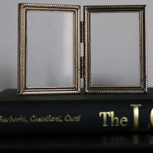 May include: A hinged, silver-toned picture frame with two rectangular openings. The frames have a rope-like border and are connected by a hinge. The frame is resting on a dark book. The overall aesthetic is vintage and elegant.