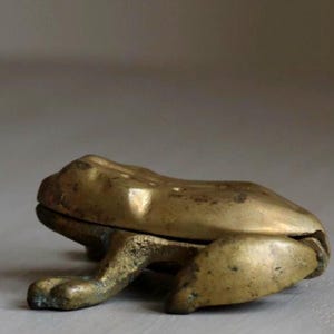 May include: A brass frog-shaped nutcracker. The frog is a golden color with a slightly aged patina. The nutcracker has a hinged mouth and detailed legs, giving it a realistic appearance. The frog is a decorative and functional item.