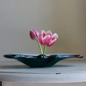 May include: A dark green, oblong-shaped vase holds two pink and white tulips. The vase has a glossy finish and a dark red rim. The tulips are in full bloom, with their petals displaying a gradient of pink and white.