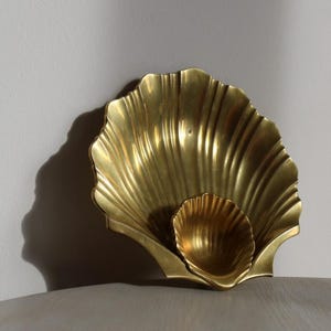 May include: A golden brass seashell-shaped decorative bowl. The bowl has a large, fluted outer shell and a smaller, nested inner shell. The surface reflects light, creating highlights and shadows. The bowl sits on a neutral surface.