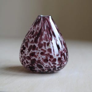 May include: A small, teardrop-shaped glass vase with a speckled pattern. The vase features a deep purple color with white and lighter purple spots. The opening is narrow, and the vase has a smooth, glossy finish. A decorative item.