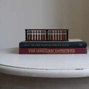 May include: A vintage wooden abacus with brown beads rests atop two books. The top book is dark blue with the title "-100 MOST BEAUTIFUL ROOMS IN AMERICA." The bottom book is burgundy with the title "THE UNICORN TAPESTRIES."