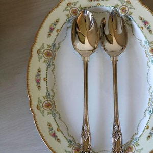 May include: A silver serving spoon and fork rest on a white oval plate with a floral border. The plate has a scalloped edge and a gold trim. The serving utensils have ornate handles.