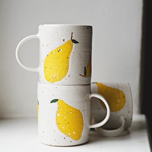 Peut inclure: Trois mugs en céramique blanche empilés, chacun orné d'une grande illustration de poire jaune. Les poires ont des feuilles vertes et des tiges marron. Les mugs ont une texture mouchetée et une anse incurvée.