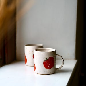 Op de afbeelding: Twee cilindrische keramische mokken met een gespikkelde gebroken witte afwerking, elk voorzien van een handgeschilderd rood tomatenontwerp met een groene steel. De mokken hebben een gladde rand, een stevig zijhandvat en een licht textuur.