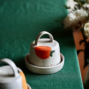 Puede incluir: Un recipiente de mantequilla de cerámica blanca con tapa y un pequeño diseño de fruta naranja. El recipiente tiene un esmalte moteado y una pequeña manija en la tapa.