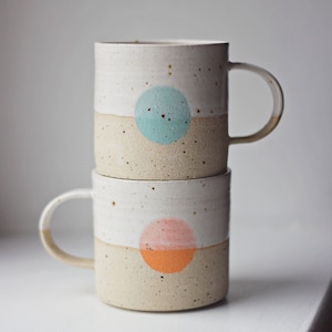 May include: Two ceramic mugs stacked on top of each other. The top mug has a light blue circle on a beige background. The bottom mug has an orange and pink circle on a beige background.