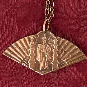 May include: A gold-toned pendant necklace with a fan-shaped design featuring a figure in a robe and a pagoda. The pendant is attached to a gold chain.