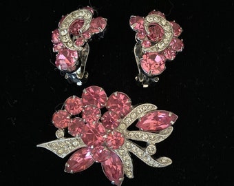 Può includere: Un set vintage di spilla e orecchini con strass rosa. La spilla presenta un design floreale con strass rosa e dettagli argentati. Gli orecchini sono di tipo clip e presentano un design simile con strass rosa.