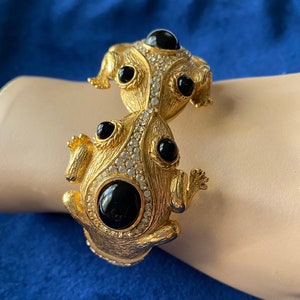 Könnte beinhalten: Ein goldfarbenes Armband mit zwei Froschfiguren mit schwarzen Onyx-Augen und klaren Strasssteinen. Das Armband ist offen und verstellbar.
