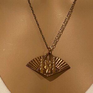 May include: A gold necklace with a pendant featuring a fan-shaped design with a figure standing in front of a temple. The pendant is made of brass and has a detailed design.