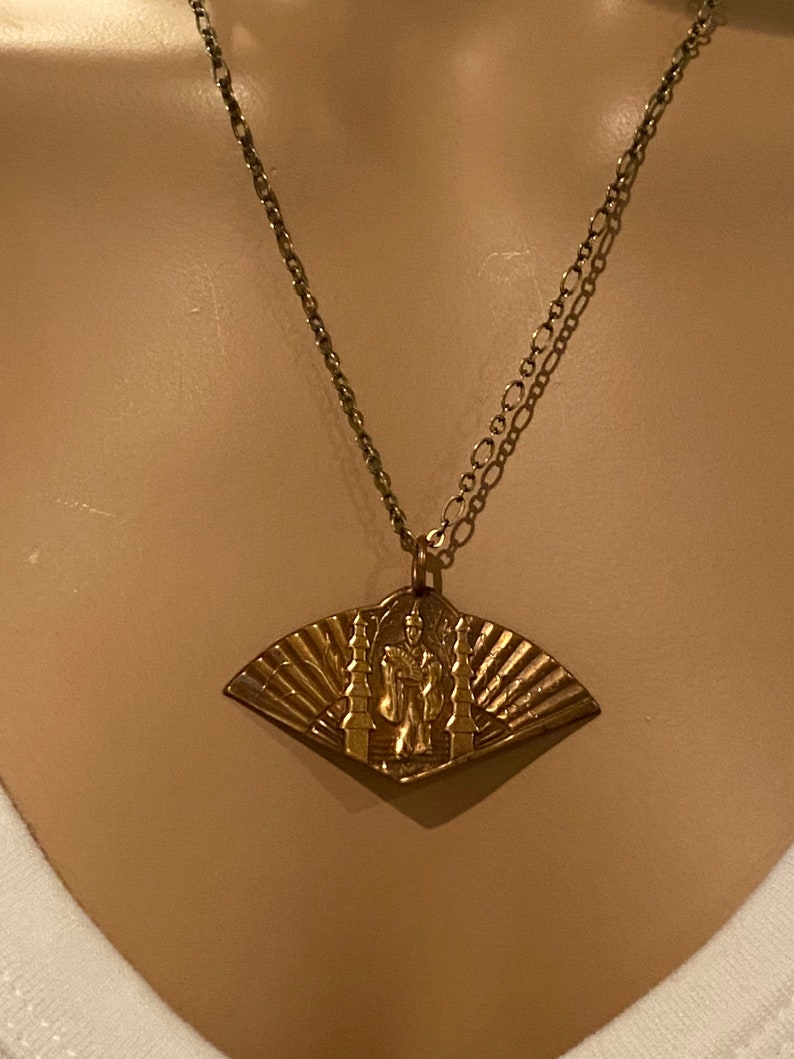 May include: A gold necklace with a pendant featuring a fan-shaped design with a figure standing in front of a temple. The pendant is made of brass and has a detailed design.