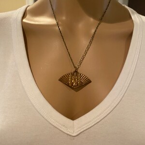 May include: A gold necklace with a fan-shaped pendant featuring a detailed design. The pendant is made of metal and has a textured surface.