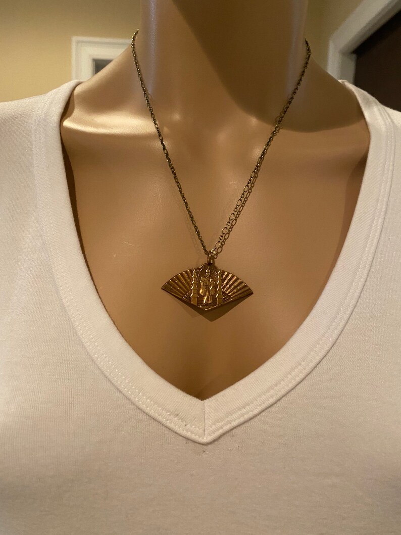 May include: A gold necklace with a fan-shaped pendant featuring a detailed design. The pendant is made of metal and has a textured surface.