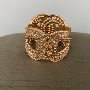 May include: A gold-toned metal bracelet with a textured, patterned design. The bracelet is open and has a rounded shape.