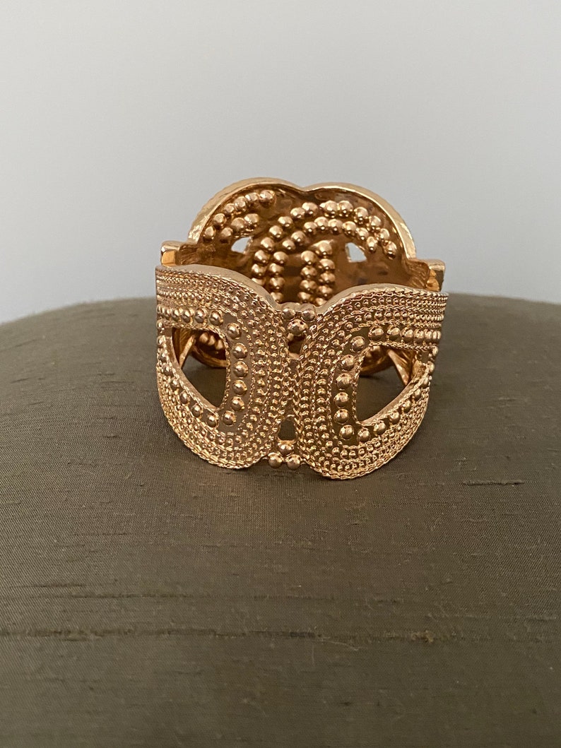 May include: A gold-toned metal bracelet with a textured, patterned design. The bracelet is open and has a rounded shape.