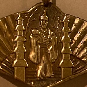 May include: A gold-colored pendant featuring a stylized figure in a robe holding a fan, flanked by two pagoda-like structures. The pendant has a textured, sunburst-like design around the edges.
