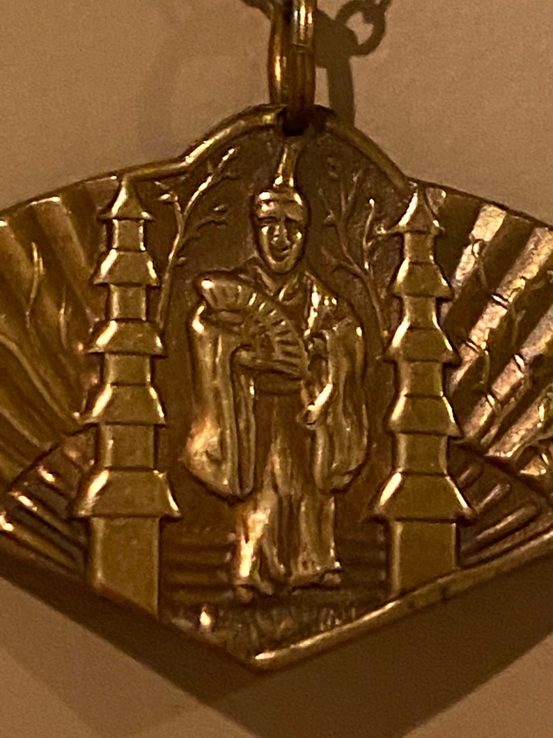 May include: A gold-colored pendant featuring a stylized figure in a robe holding a fan, flanked by two pagoda-like structures. The pendant has a textured, sunburst-like design around the edges.