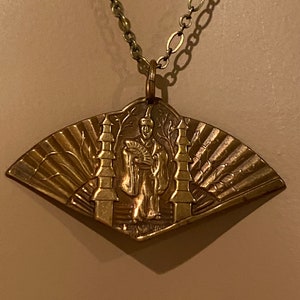 May include: A gold-toned necklace with a pendant featuring a detailed embossed design of a person standing between two towers. The pendant is shaped like a fan.
