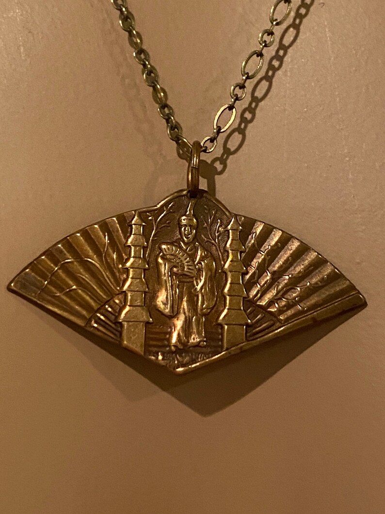 May include: A gold-toned necklace with a pendant featuring a detailed embossed design of a person standing between two towers. The pendant is shaped like a fan.