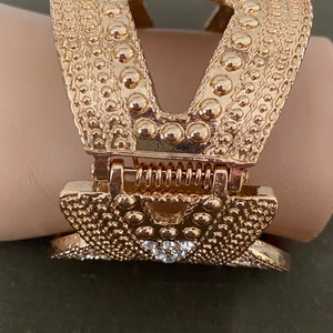 May include: Gold-toned metal hair clip with a textured surface and a decorative design. The clip has a spring mechanism and a clear rhinestone accent.