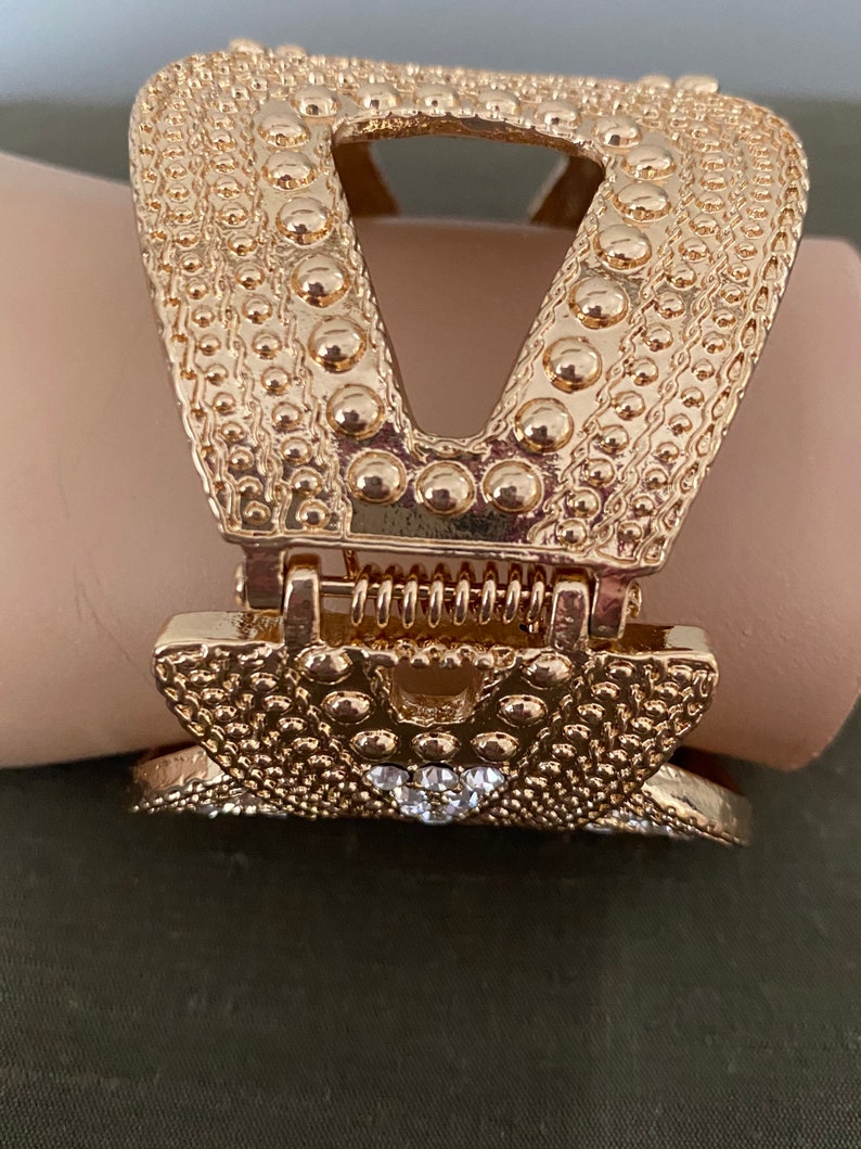 May include: Gold-toned metal hair clip with a textured surface and a decorative design. The clip has a spring mechanism and a clear rhinestone accent.