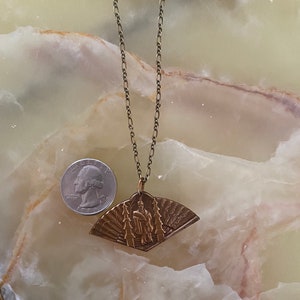 May include: A gold chain necklace with a fan-shaped pendant featuring a raised image of a person in traditional clothing. The pendant has a detailed design with intricate lines and patterns.