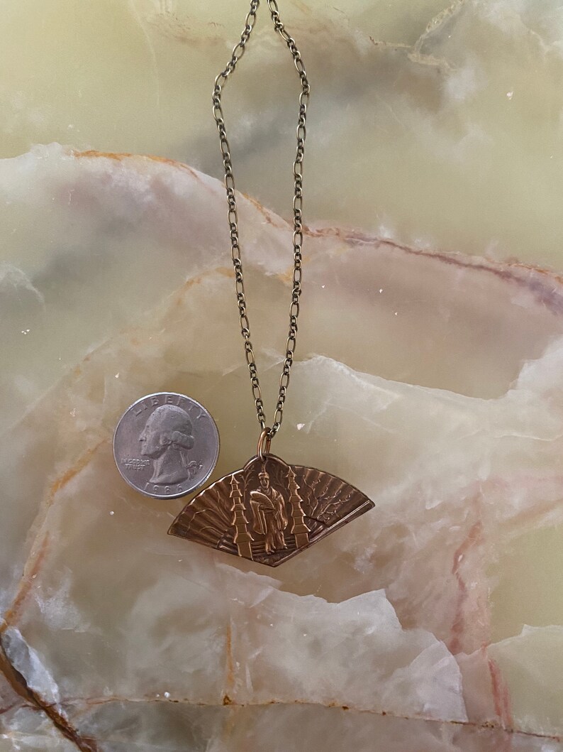 May include: A gold chain necklace with a fan-shaped pendant featuring a raised image of a person in traditional clothing. The pendant has a detailed design with intricate lines and patterns.
