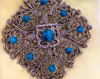 May include: A square-shaped brooch featuring intricate silver-toned filigree metalwork arranged in a floral pattern. The piece is accented with seven round, polished turquoise-colored cabochons, with one centered and six positioned at the petal points.