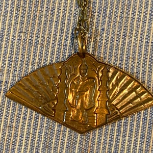 May include: A gold-toned pendant necklace with a fan-shaped design featuring a raised image of a person. The pendant is attached to a gold chain.