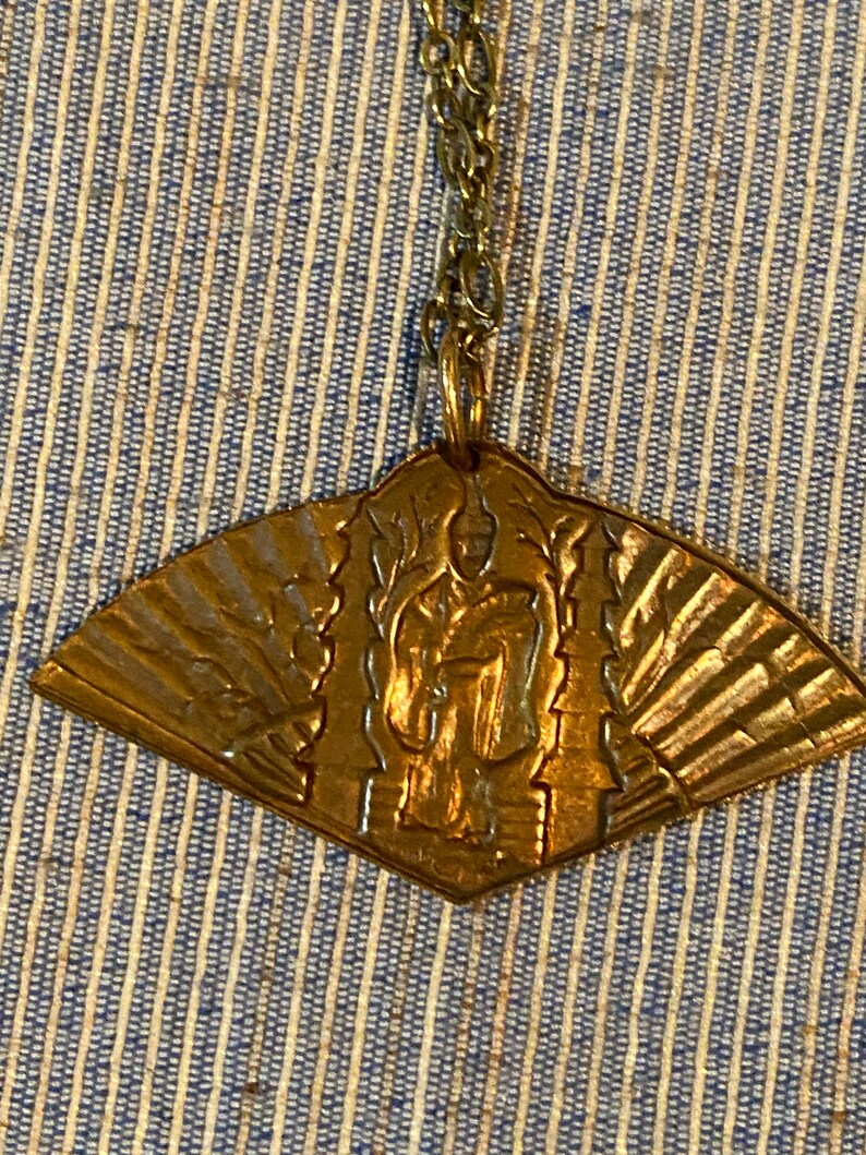 May include: A gold-toned pendant necklace with a fan-shaped design featuring a raised image of a person. The pendant is attached to a gold chain.