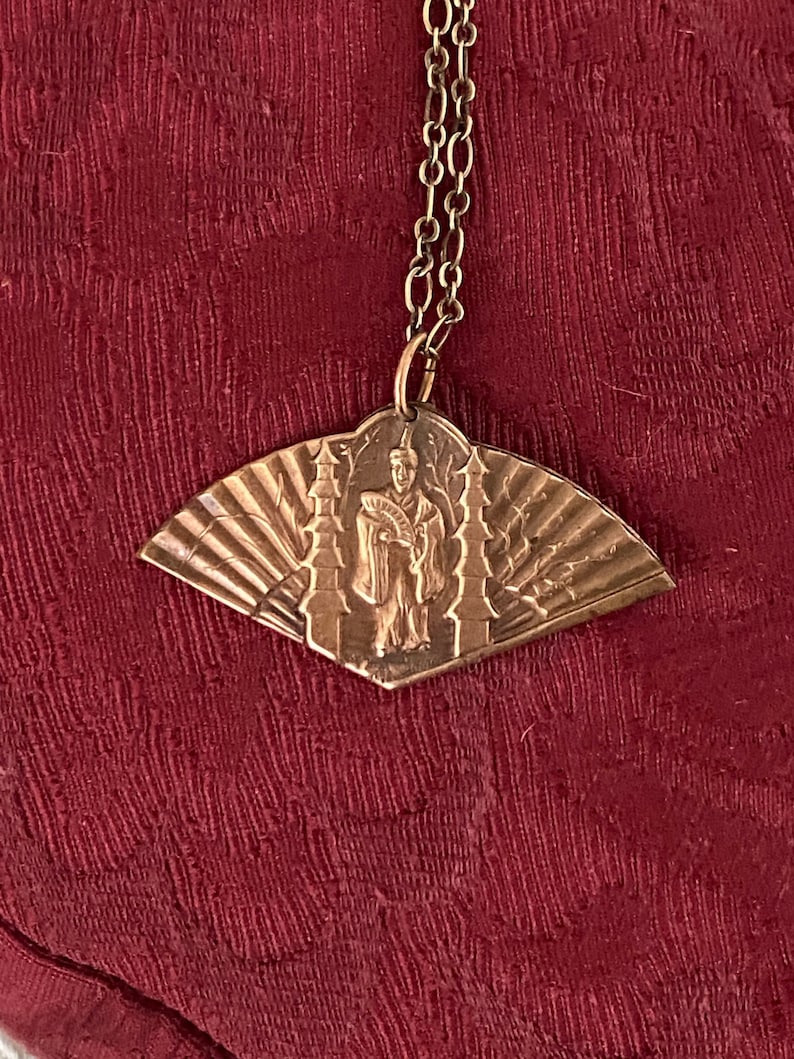 May include: A gold-toned pendant necklace with a fan-shaped design featuring a figure in a robe and a pagoda. The pendant is attached to a gold chain.