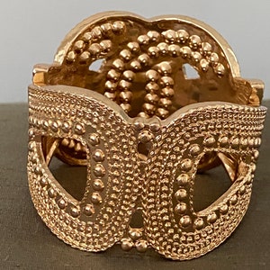 May include: A gold-toned metal bracelet with a textured, intricate design. The bracelet is open and has a hinged clasp.