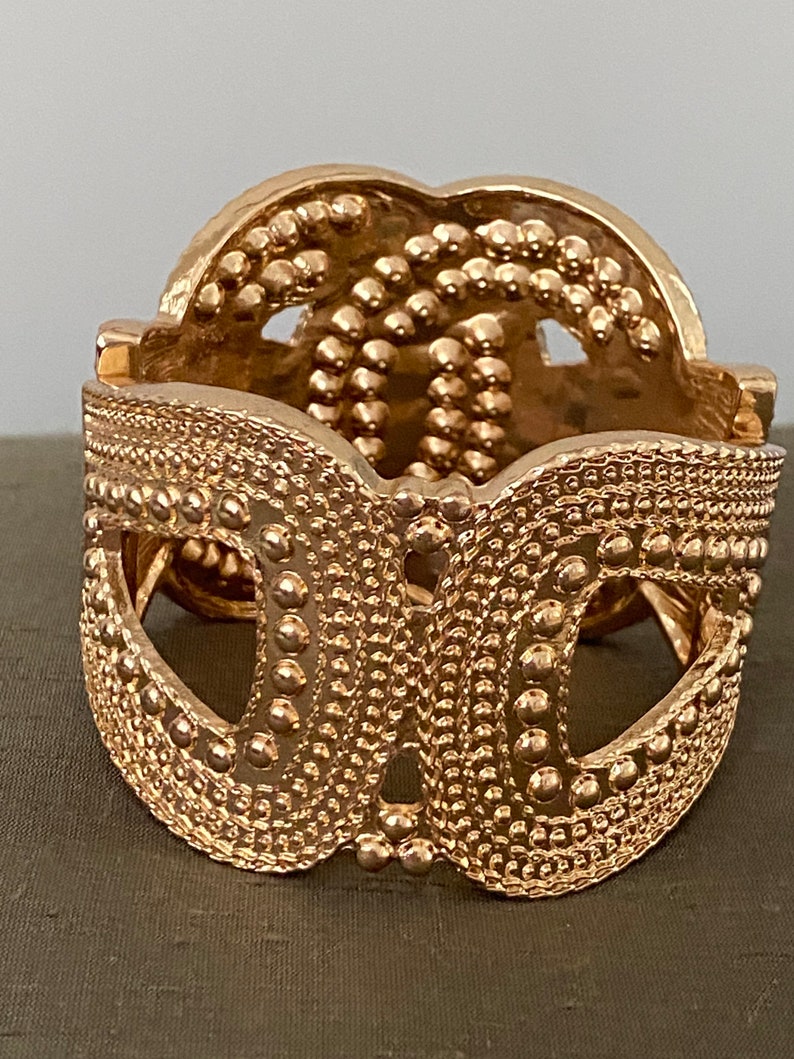 May include: A gold-toned metal bracelet with a textured, intricate design. The bracelet is open and has a hinged clasp.