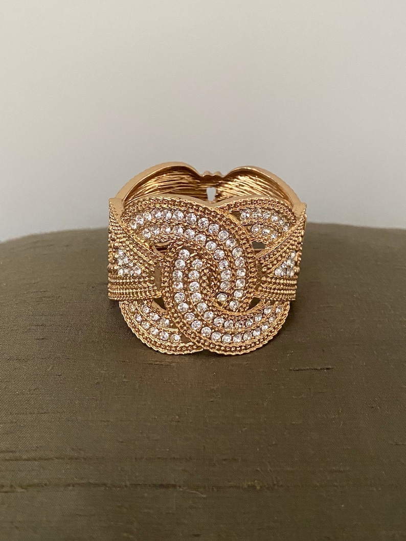 May include: A gold-toned bangle bracelet with a decorative design of interlocking circles encrusted with clear rhinestones.