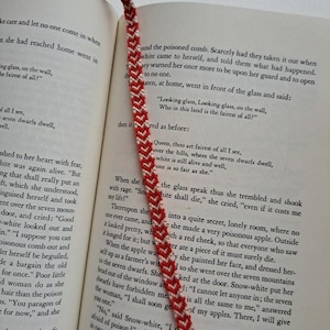 May include: A close-up of a book with a red and white friendship bracelet bookmark. The book is open to a page with text about a poisoned apple and a queen. The text reads, "Queen, thou art fairest of all I see, Over the hills, where the seven dwarfs dwell, White is still alive and well, One is so fair as she."