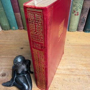 May include: A red hardback book titled Brontë's Works Jane Eyre with gold embossed spine detailing, positioned next to a small black figurine of a kneeling person. The book features a textured red cover and gold lettering on the spine.