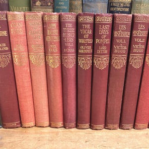 May include: A collection of antique books with red and gold covers, including titles like "The Moonstone" and "Les Miserables." A bronze-colored sculpture of a woman sits to the left of the books. The books are arranged on a wooden surface.