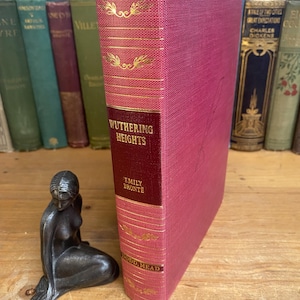 May include: A vintage copy of "Wuthering Heights" by Emily Brontë, bound in a deep rose-colored cloth with gold lettering and accents. The book is displayed next to a small, dark-toned sculpture of a seated woman.
