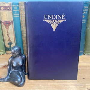 May include: A dark blue book titled "UNDINE" with gold lettering and a decorative emblem. A small, silver mermaid statue sits in front of the book. Other antique books are visible in the background, with titles like "Villette" and "Jane Eyre".